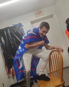 Bobby in Todd Rhode’s shop, draped in a Trump flag to imitate Ashli Babbitt.